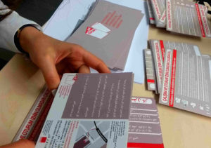 UNDOK-Flyer und Info-Postkaften auf verschiedenen Sprachen auf einem Tisch verteilt. Zwei Hände, die eine Postkoarte halten.